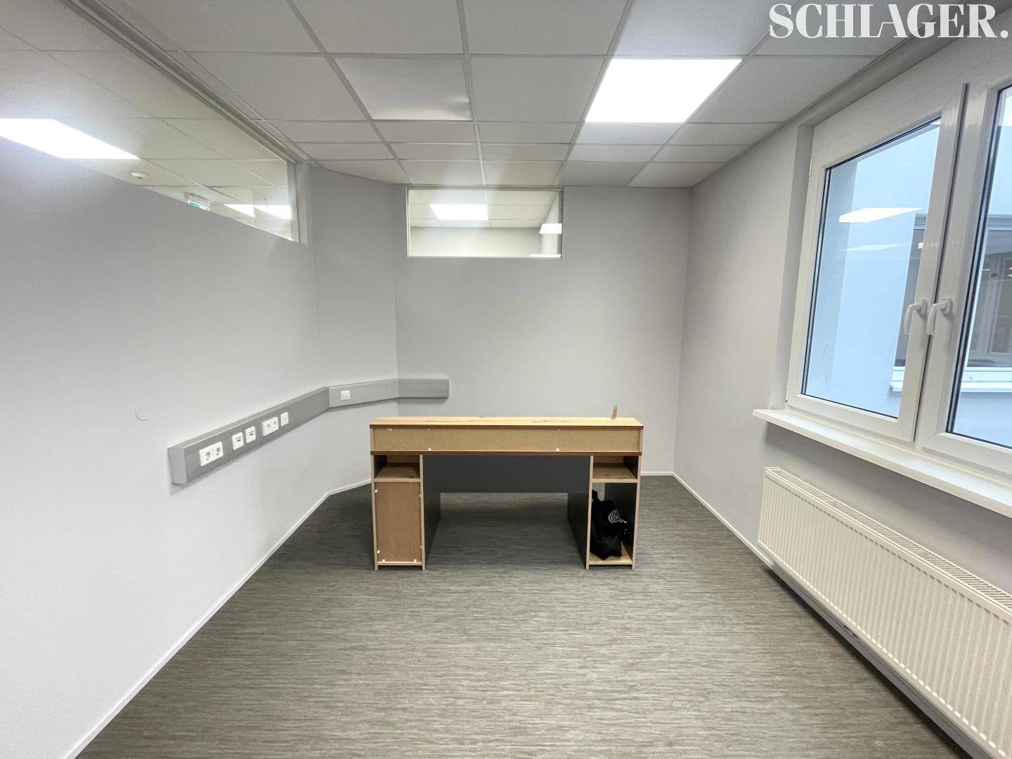 Kleineres Büro mit einem einfachen Schreibtisch, Fenster und praktischen Wandanschlüssen.