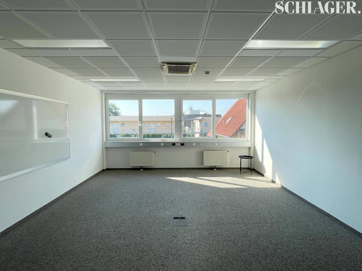 Heller Büroraum mit Teppichboden, großen Fenstern und einer weißen Tafel an der Wand.