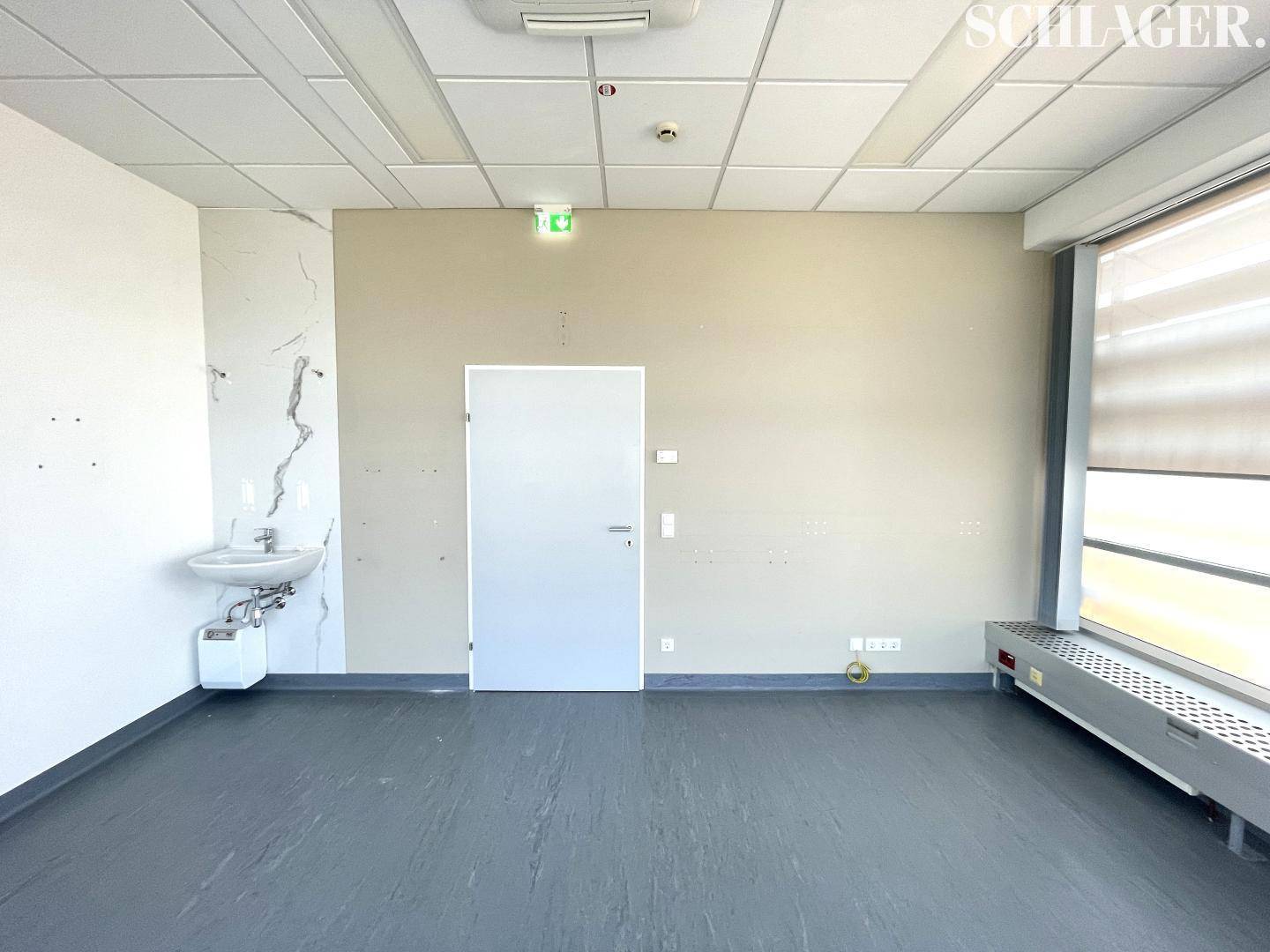 Heller Büroraum mit einem Waschbecken an der Wand und einer Tür, die zu weiteren Räumen führt.