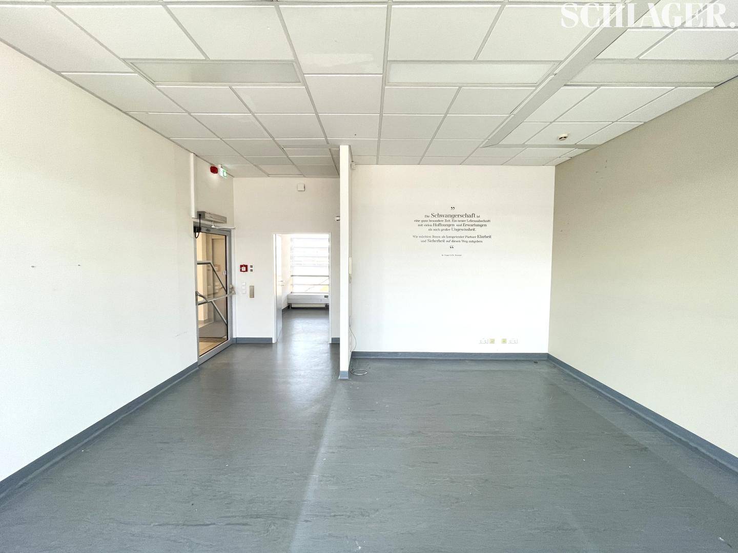 Offener Bürobereich mit einer Trennwand und einem Ausgang, der zu einem weiteren Flur führt.