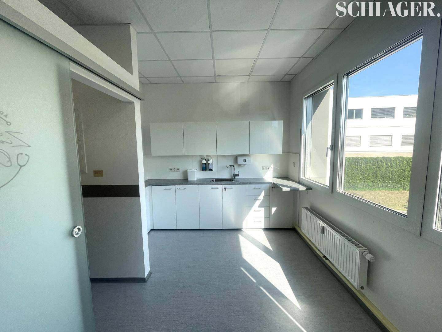 Helle Teeküche mit weißen Unterschränken, Spülbecken und einem Heizkörper unter dem großen Fenster.