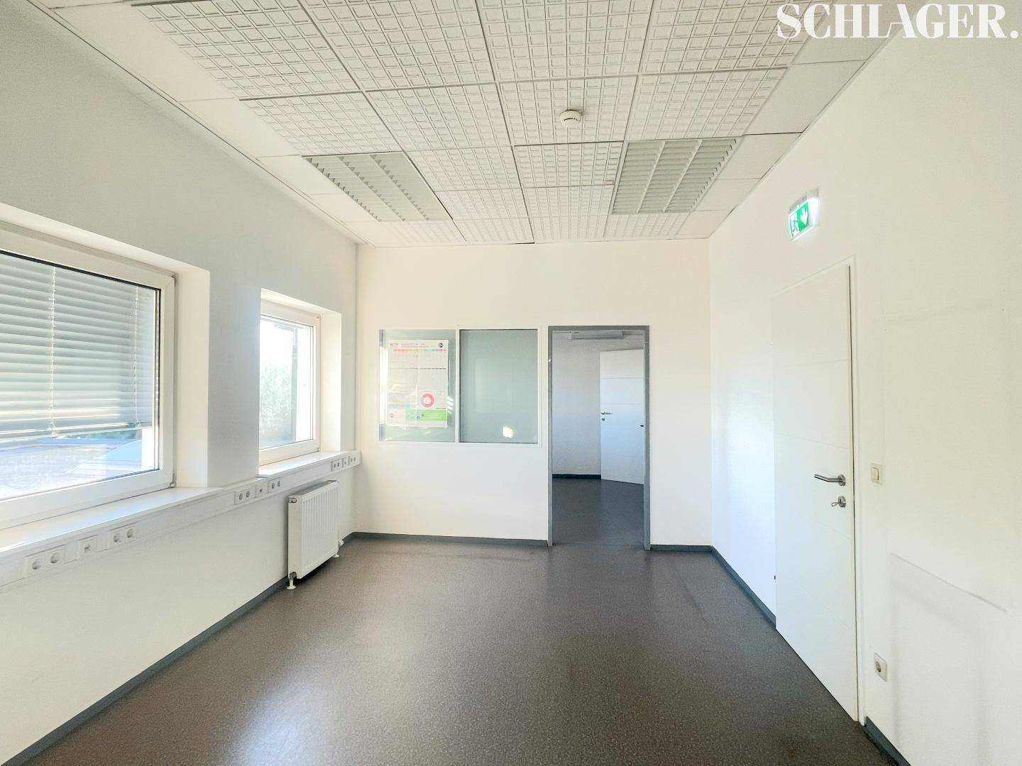 Funktionaler Büroraum mit Teppichboden, Fenstern und Zugang zu weiteren Bereichen, ideal für Arbeitsplätze.