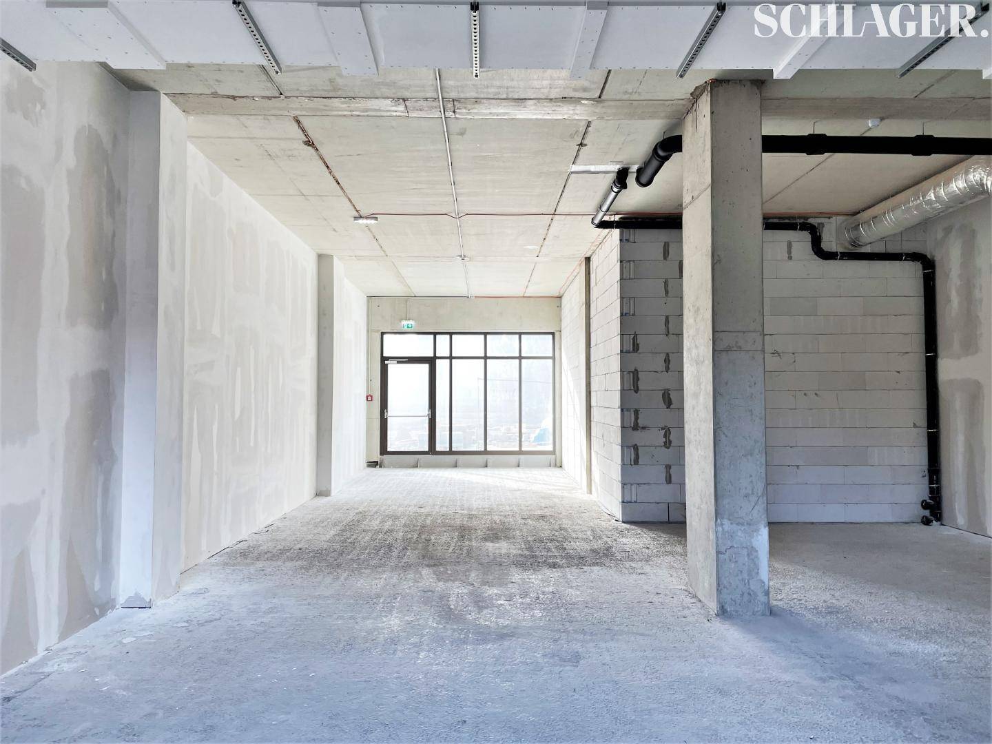 Unfertiger Innenraum mit Betonboden, sichtbaren Leitungen und einem großen Fenster am Ende des Raumes.