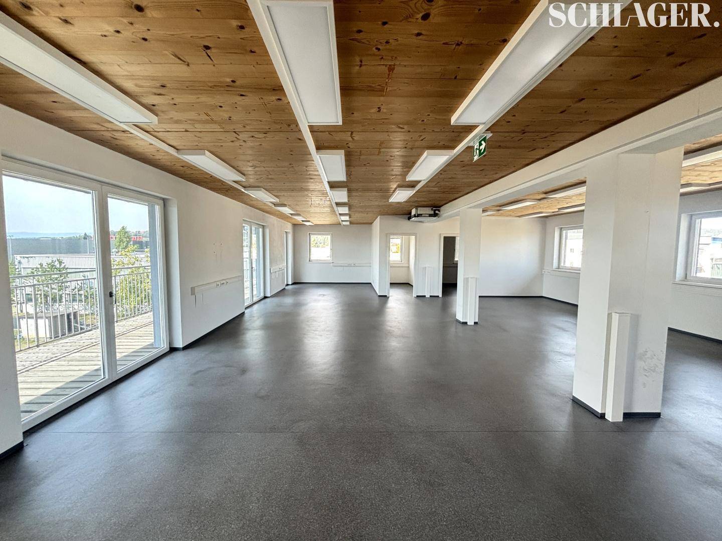 Heller Büroraum mit mehreren Balkonzugängen, Holzbalkendecke und glattem Betonboden.