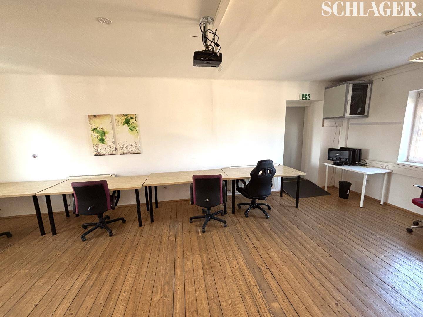 Arbeitsraum mit Holzdielenboden, mehreren Tischen, Bürostühlen und einer Klimaanlage an der Wand.