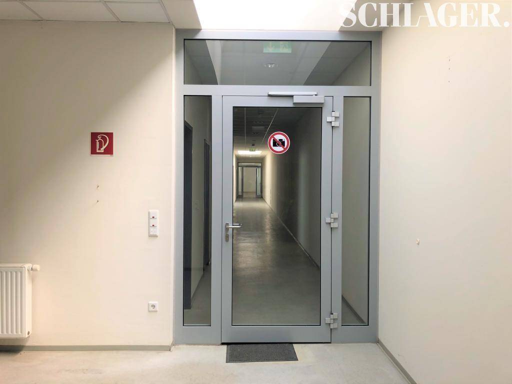 Eingangsbereich zu einem langen Flur mit moderner Glastür und Brandschutzzeichen an der Wand.