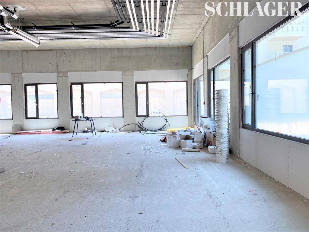 Unfertiger Innenraum mit Betonboden, sichtbaren Deckeninstallationen und mehreren Fenstern, Baumaterialien sind zu sehen.