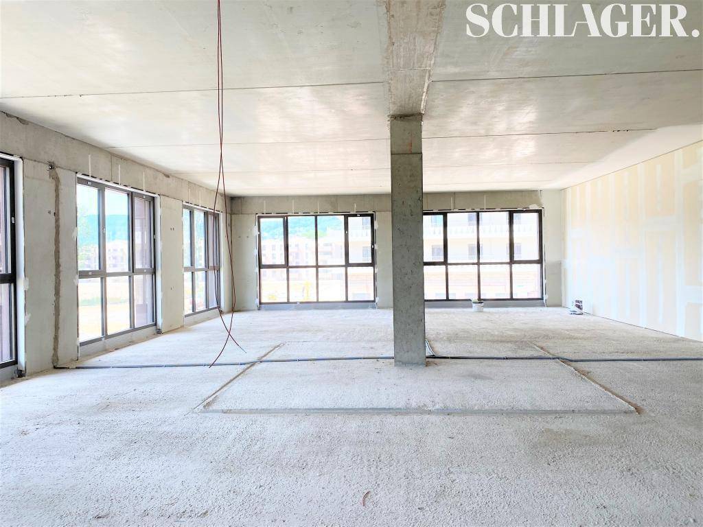 Großer, unfertiger Innenraum mit Betonboden und -decke, einem Pfeiler und vielen großen Fenstern, im Rohbauzustand.