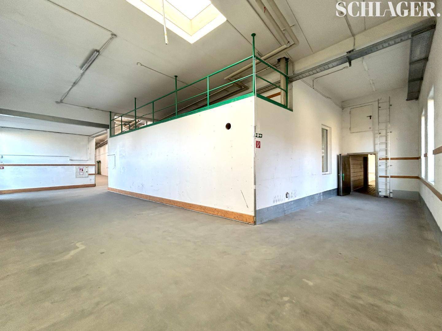Großer Lagerraum mit Betonboden, hoher Decke und einer erhöhten Plattform mit grünem Geländer.