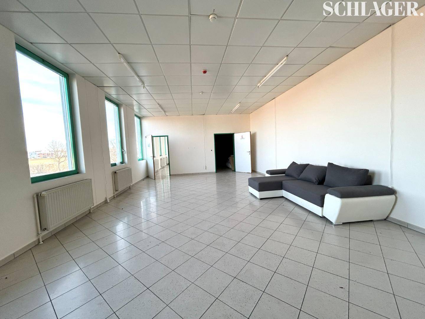 Heller Büroraum mit Fliesenboden, großen Fenstern und einem modernen Ecksofa in Grau-Weiß.