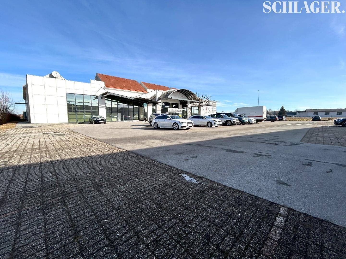Gesamtansicht der Gewerbeimmobilie mit weitläufigem Parkplatz und mehreren Fahrzeugen unter blauem Himmel.