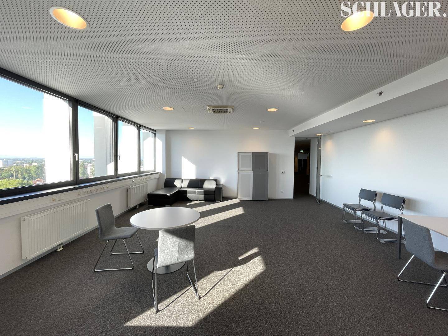 Offener Bürobereich mit Sitzgelegenheiten, Kühlschrank und viel Tageslicht durch große Fenster.