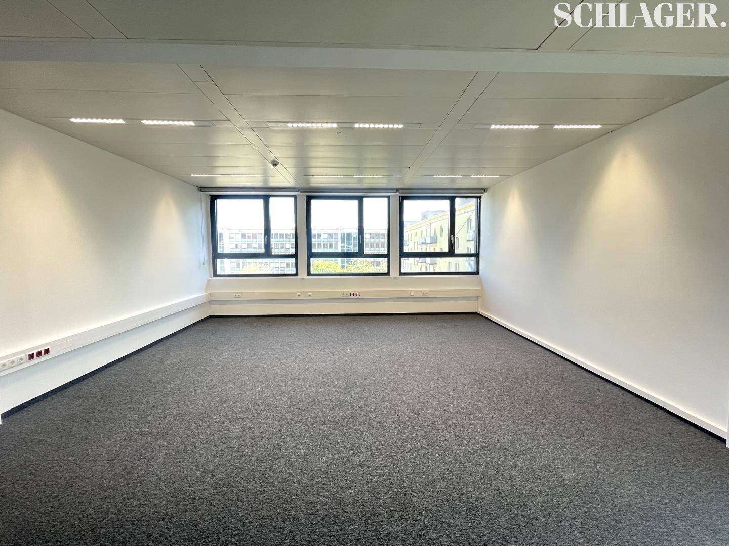 Geräumiges Büro mit Teppichboden, einer Fensterfront und moderner Deckenbeleuchtung.