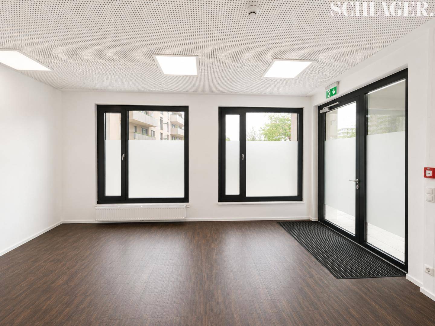 Eingangsbereich mit großen Fenstern, Milchglas im unteren Bereich, dunklem Laminatboden und weißen Wänden.
