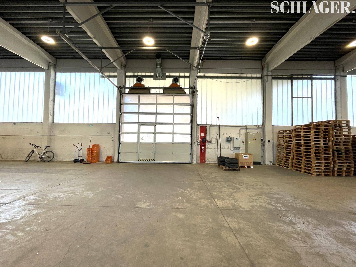 Innenansicht einer Lagerhalle mit großem Rolltor, hohen Fenstern und Betonboden für industrielle Nutzung.