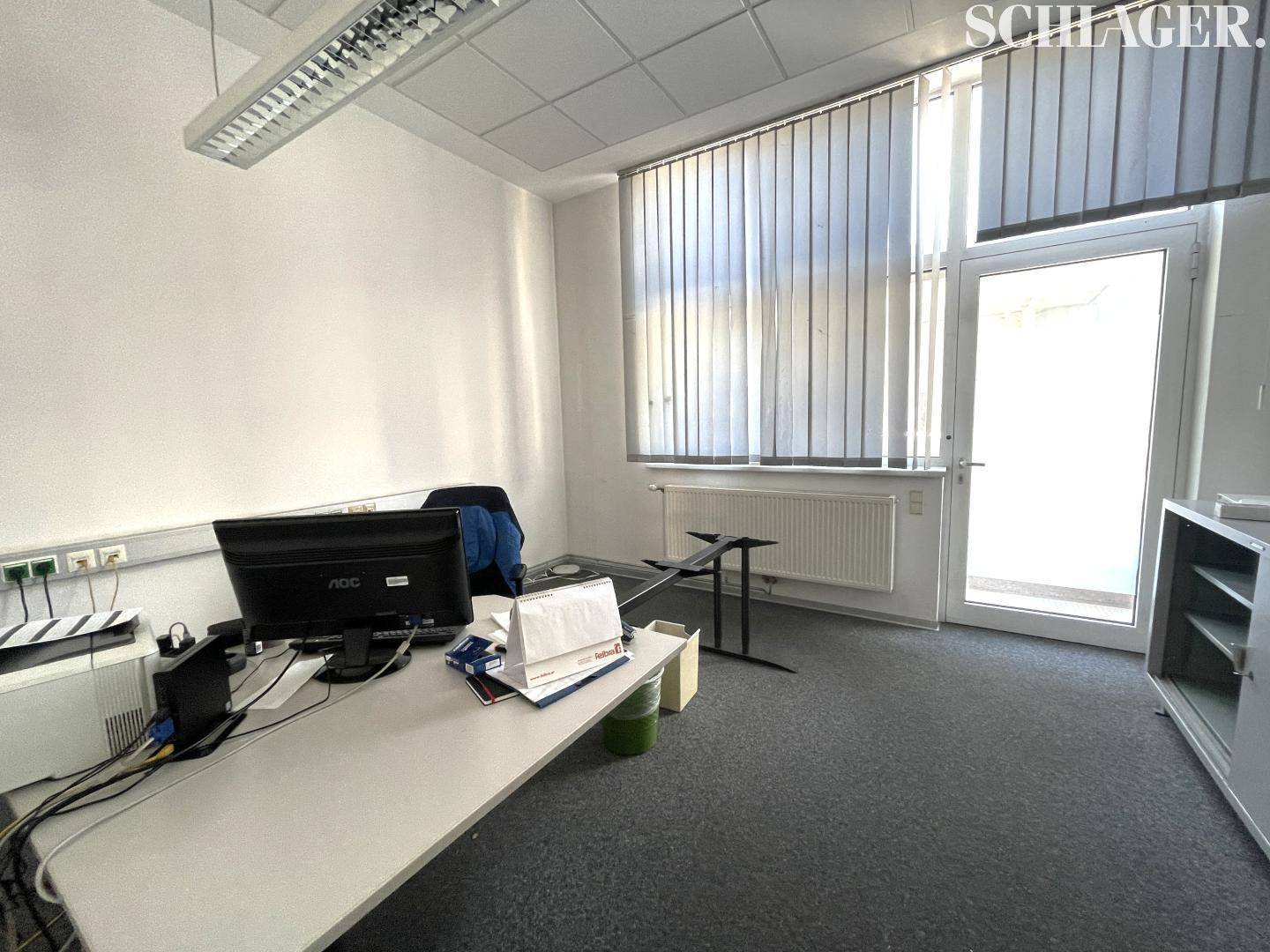 Funktionaler Büroraum mit Schreibtisch, Computer und einem Fenster mit Jalousien für natürliches Licht.
