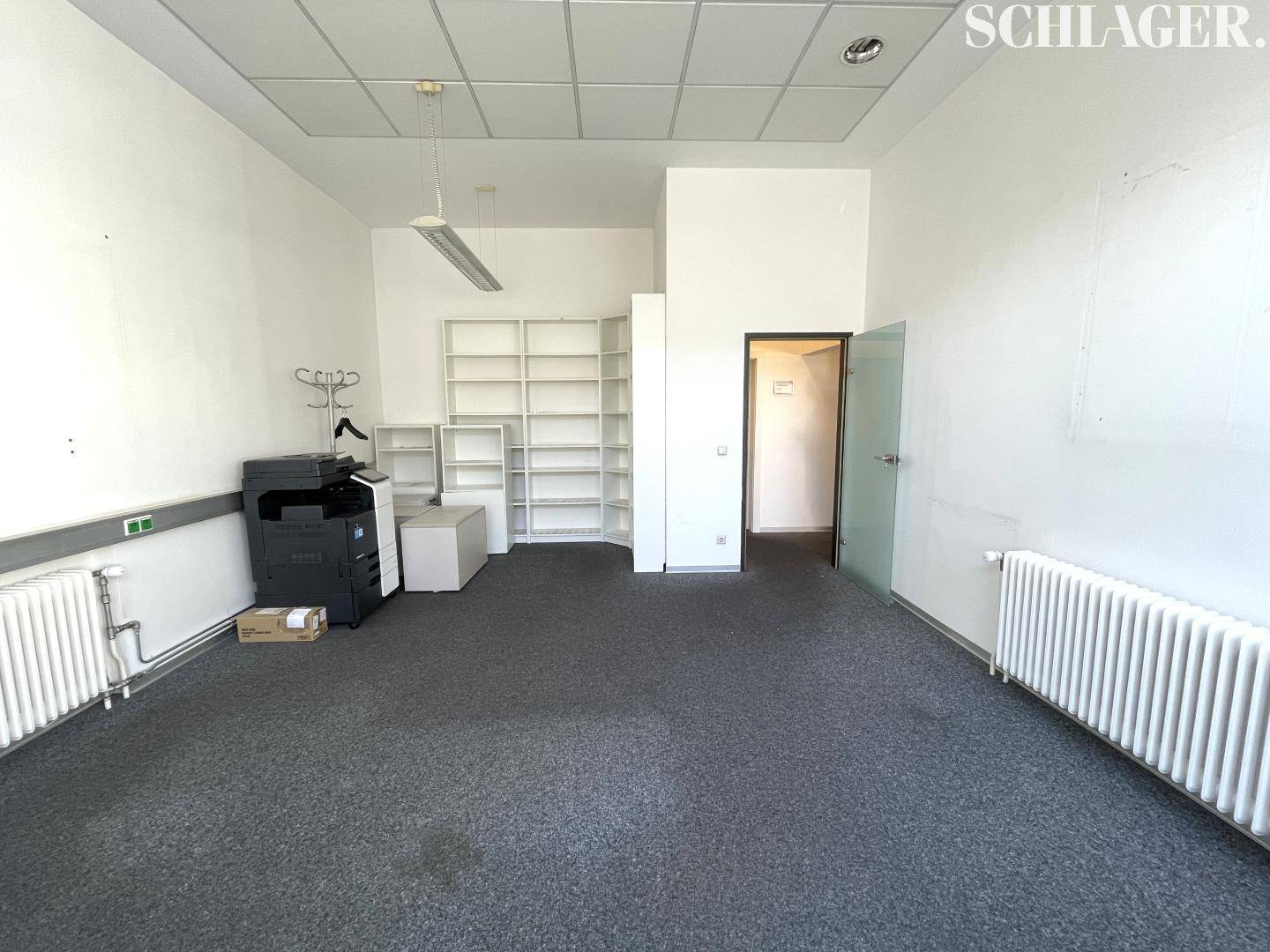 Heller Büroraum mit Teppichboden, weißen Wänden, praktischen Regalsystemen und einem Drucker.