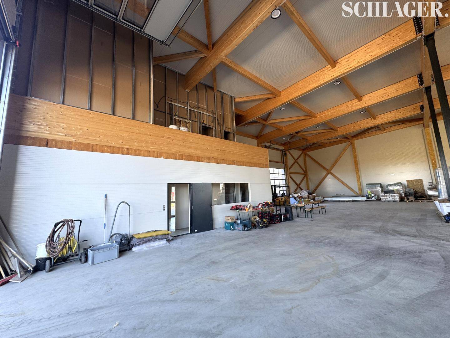 Weitläufige Industriehalle mit Betonboden, Holzkonstruktion und einem abgetrennten Bereich, der als Büro dienen könnte.