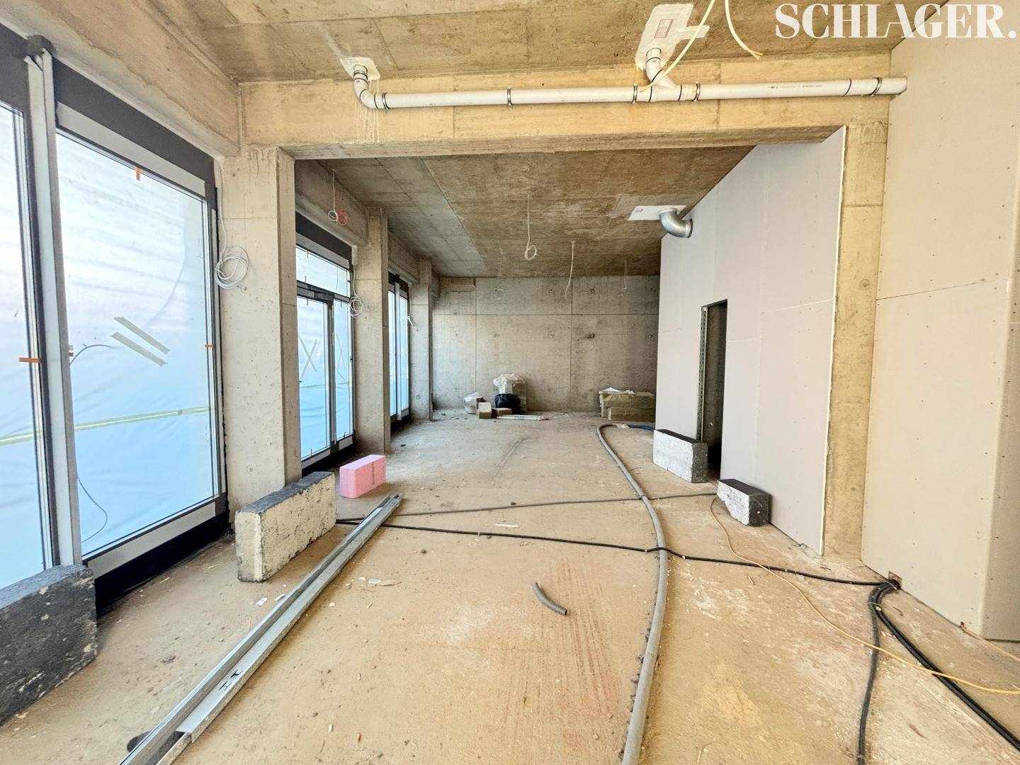 Innenraum im Rohbau mit großen Fenstern, Betonwänden und sichtbaren Installationen während der Bauphase.