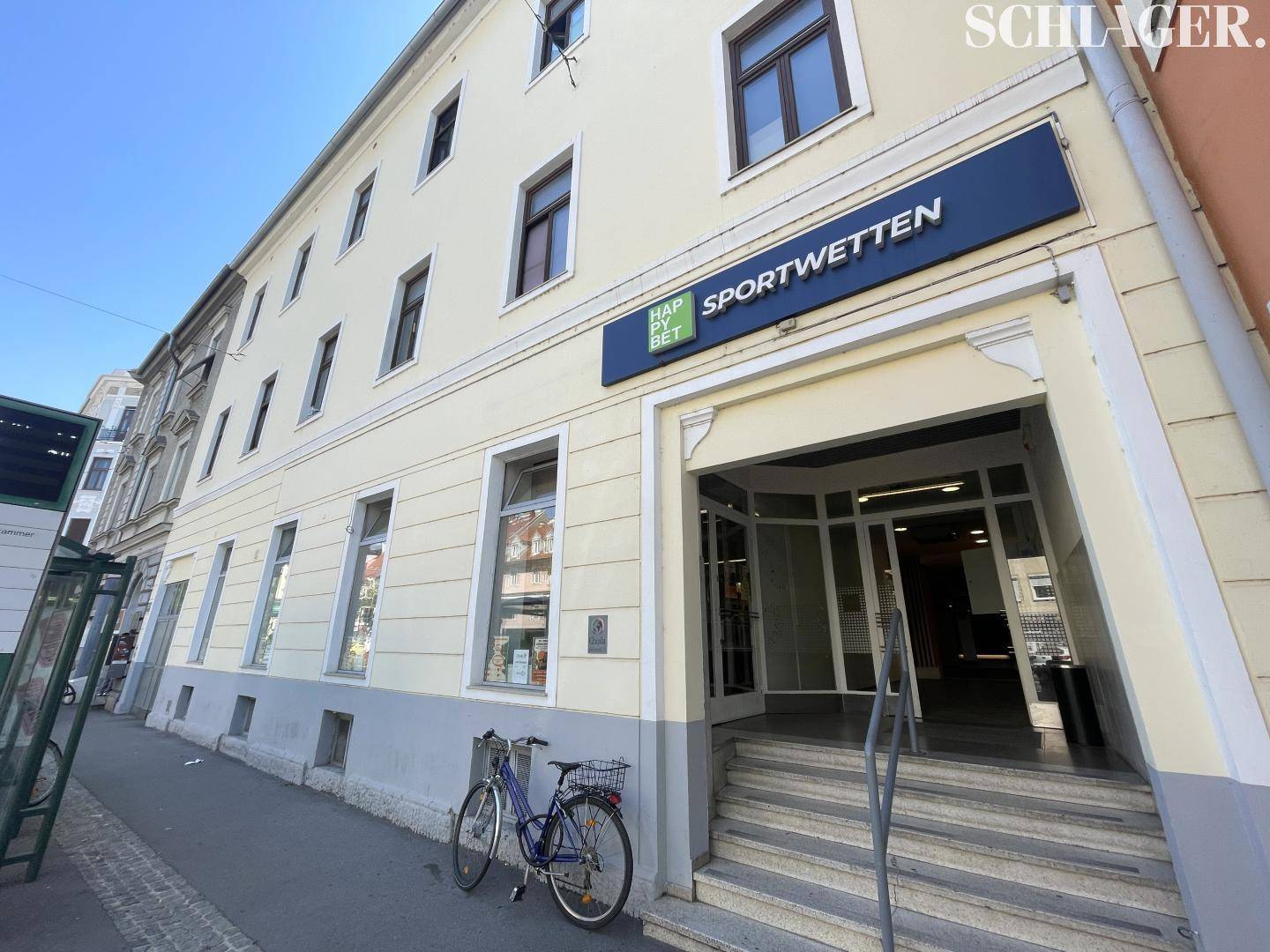 Seitenansicht des Gebäudes mit Eingangstreppe und dem "SPORTWETTEN"-Schild, ein Fahrrad steht davor.