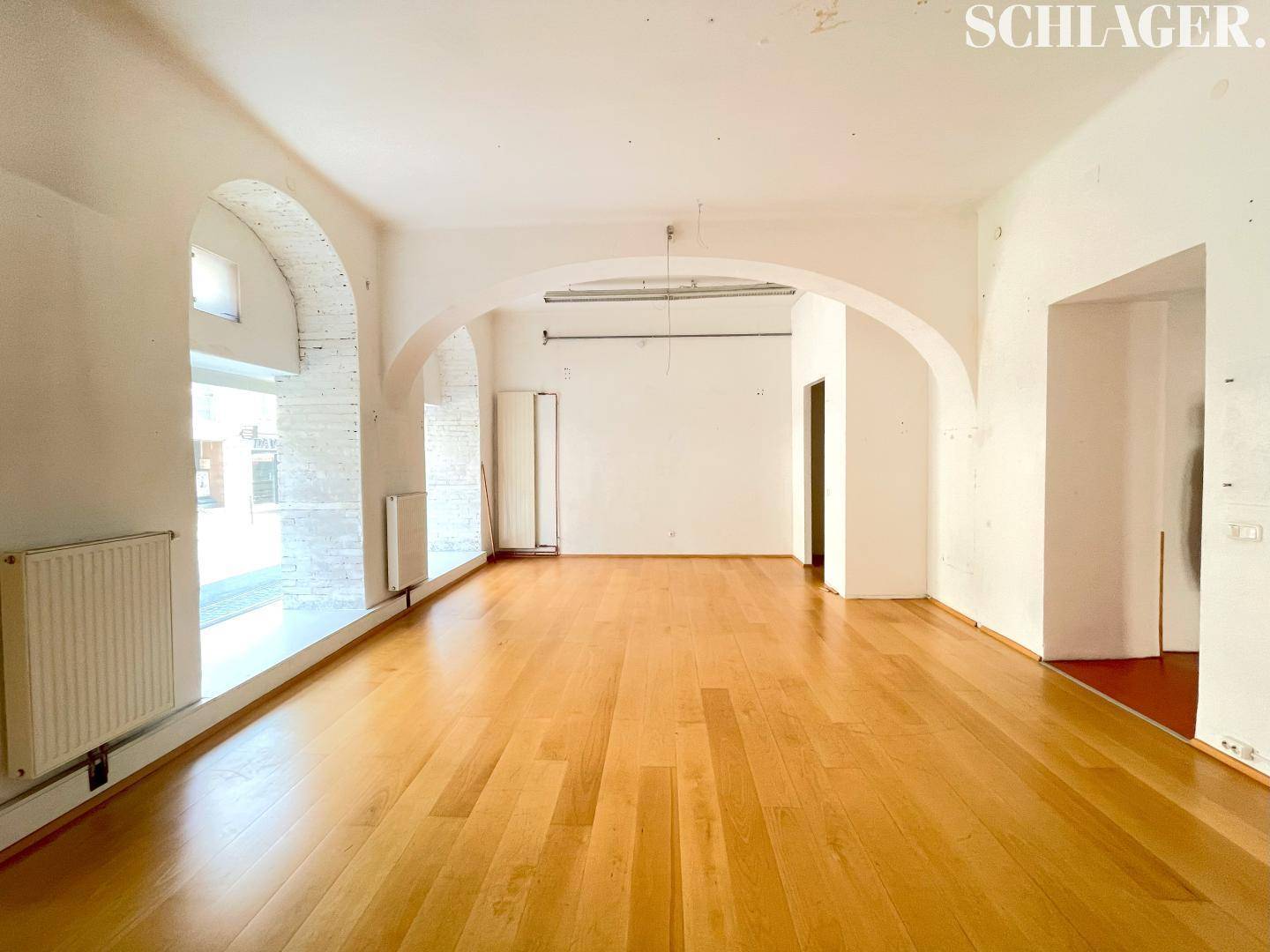 Heller Ladenbereich mit Holzboden, großen Fenstern und einem eleganten Bogen, ausgestattet mit Heizkörpern.