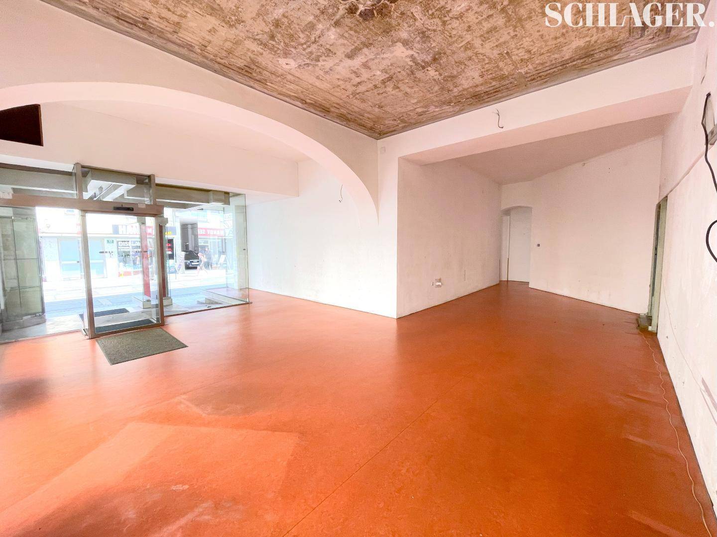 Offener Gewerberaum mit orangefarbenem Boden, großen Schaufenstern und einem modernen Glaseingangsbereich.