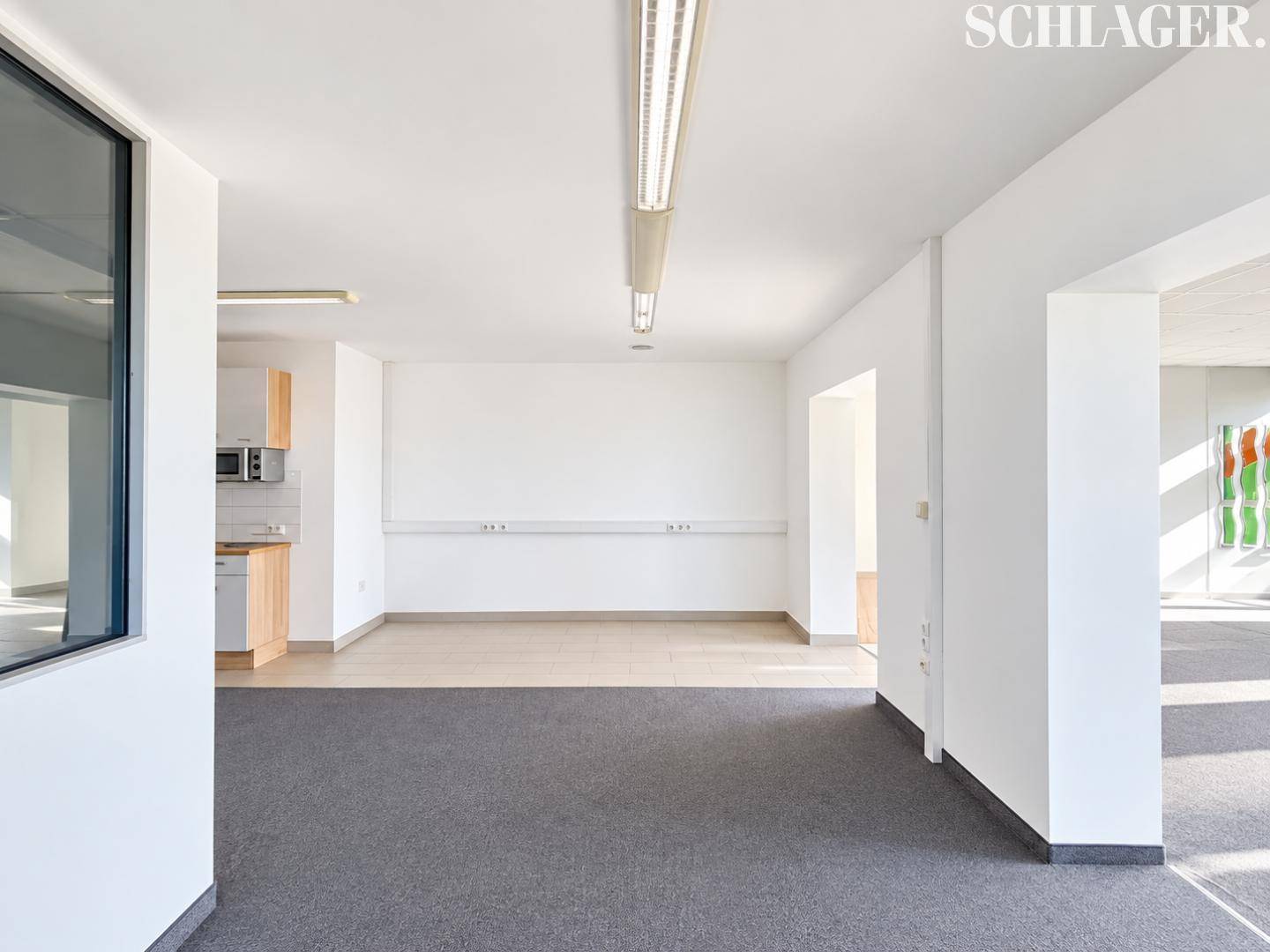 Offener Bürobereich mit Teppichboden und einer kleinen Küchenzeile im Hintergrund, gut beleuchtet.