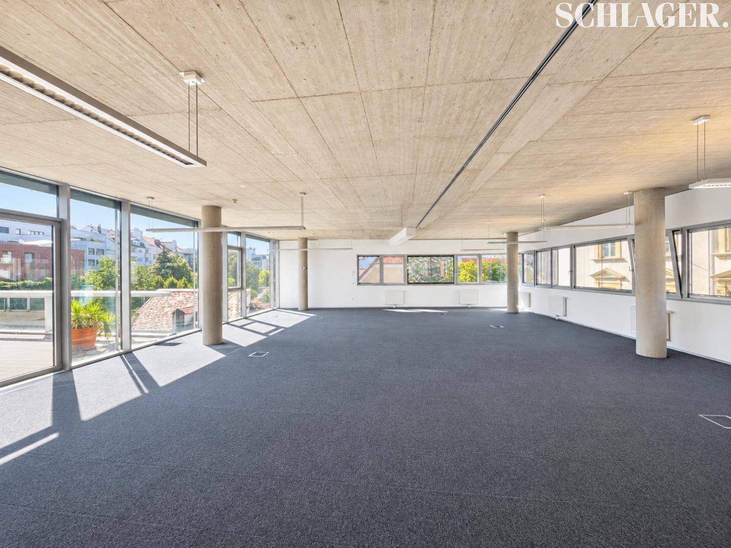 Geräumiger, offener Bürobereich mit dunklem Teppichboden, Sichtbetondecke und großen Fenstern für viel Tageslicht.
