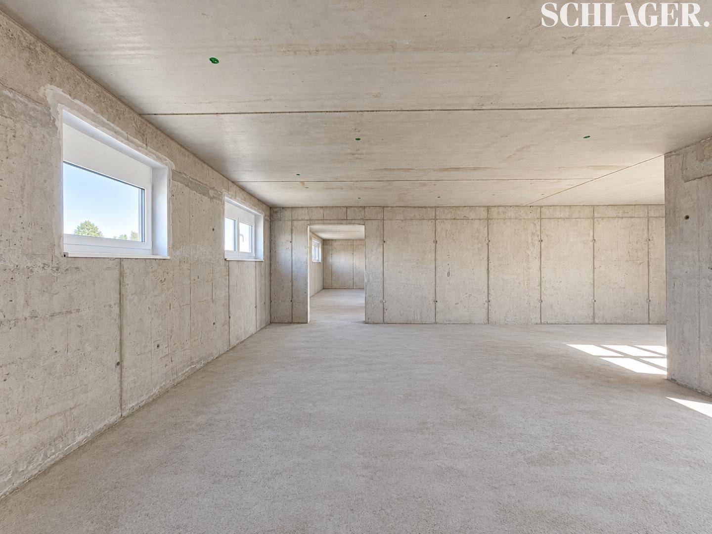 Geräumiger Rohbau-Raum mit Betonwänden und -boden, mehreren Fenstern und viel Tageslicht.