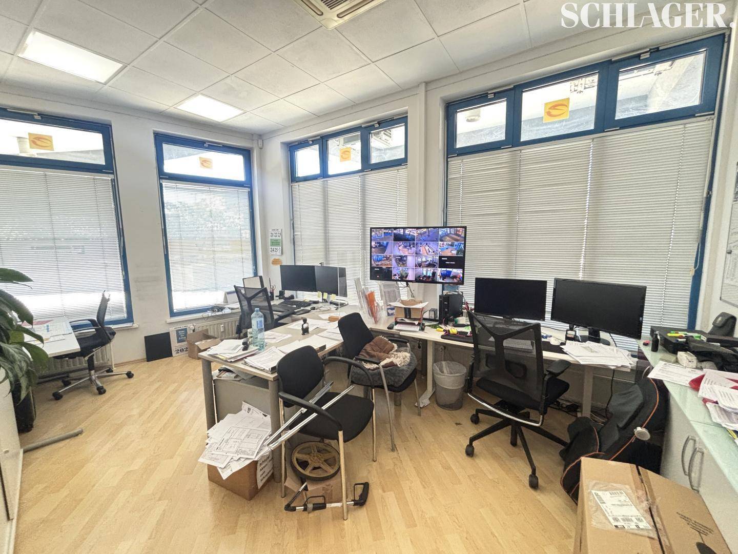 Großraumbüro mit mehreren Arbeitsplätzen, Computern und großen Fenstern mit Jalousien.