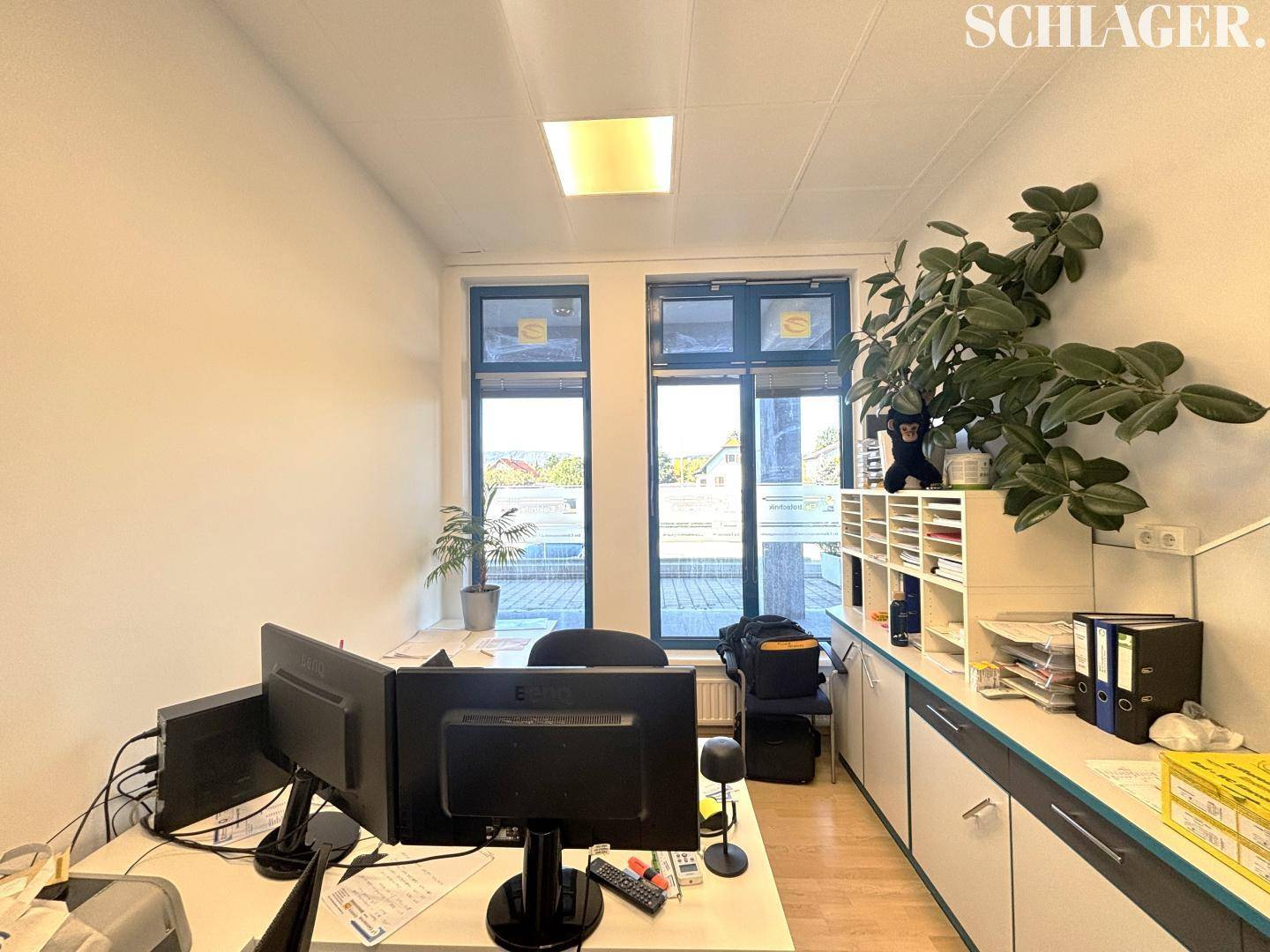 Einzelbüro mit einem Schreibtisch, Computermonitoren, einer Pflanze und hohen Fenstern mit Ausblick.