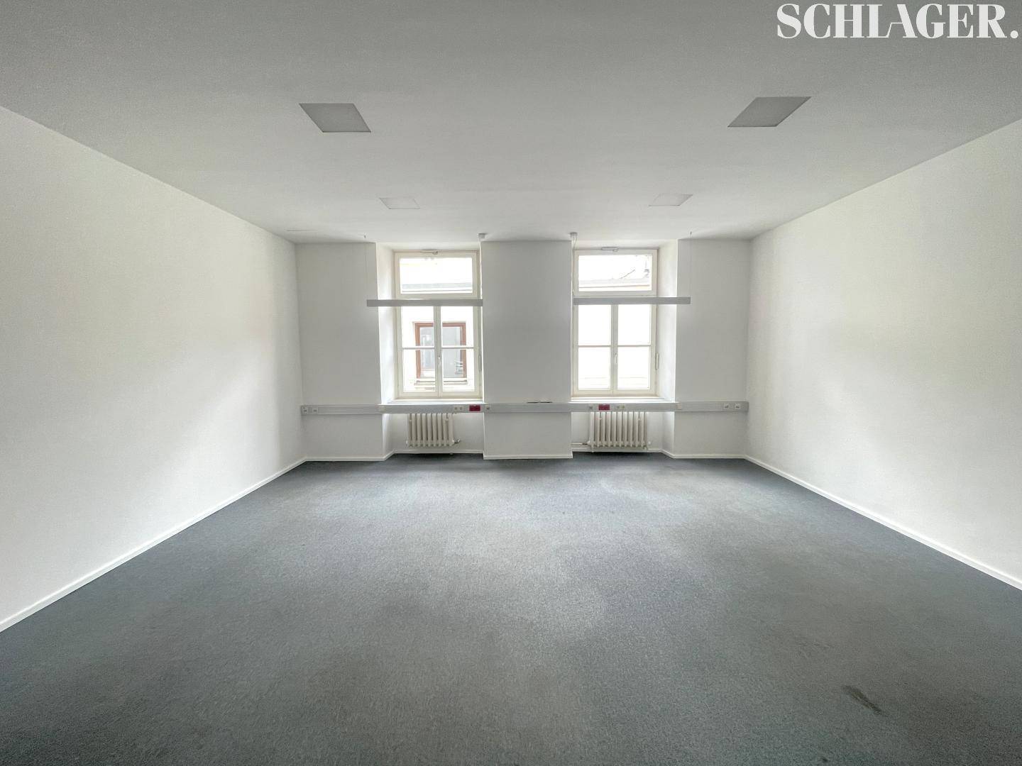 Geräumiger, leerer Büroraum mit hellem Anstrich, grauem Teppich und zwei Fenstern mit Blick ins Grüne.