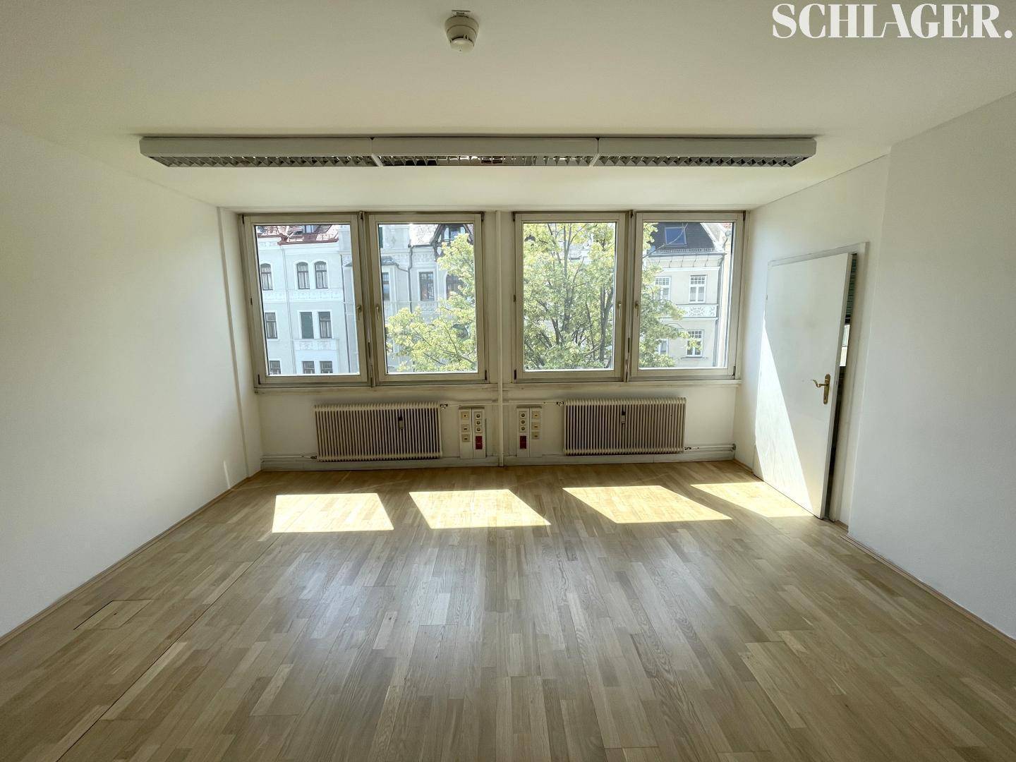 Geräumiger Büroraum mit großen Fenstern, die viel Tageslicht hereinlassen, und hellem Parkettboden.