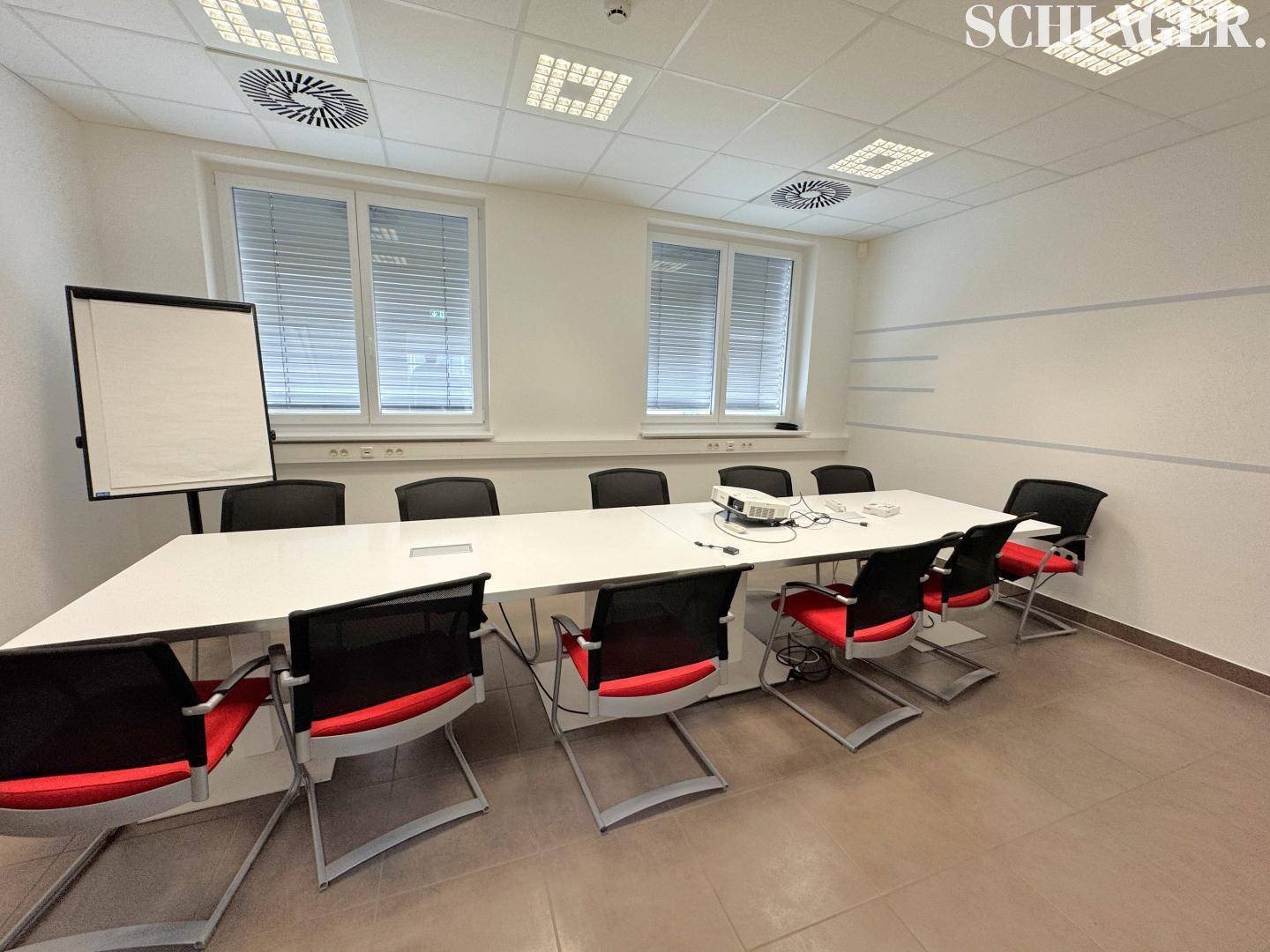 Moderner Konferenzraum mit großem Tisch, Bürostühlen, Beamer und Fenstern mit Jalousien.