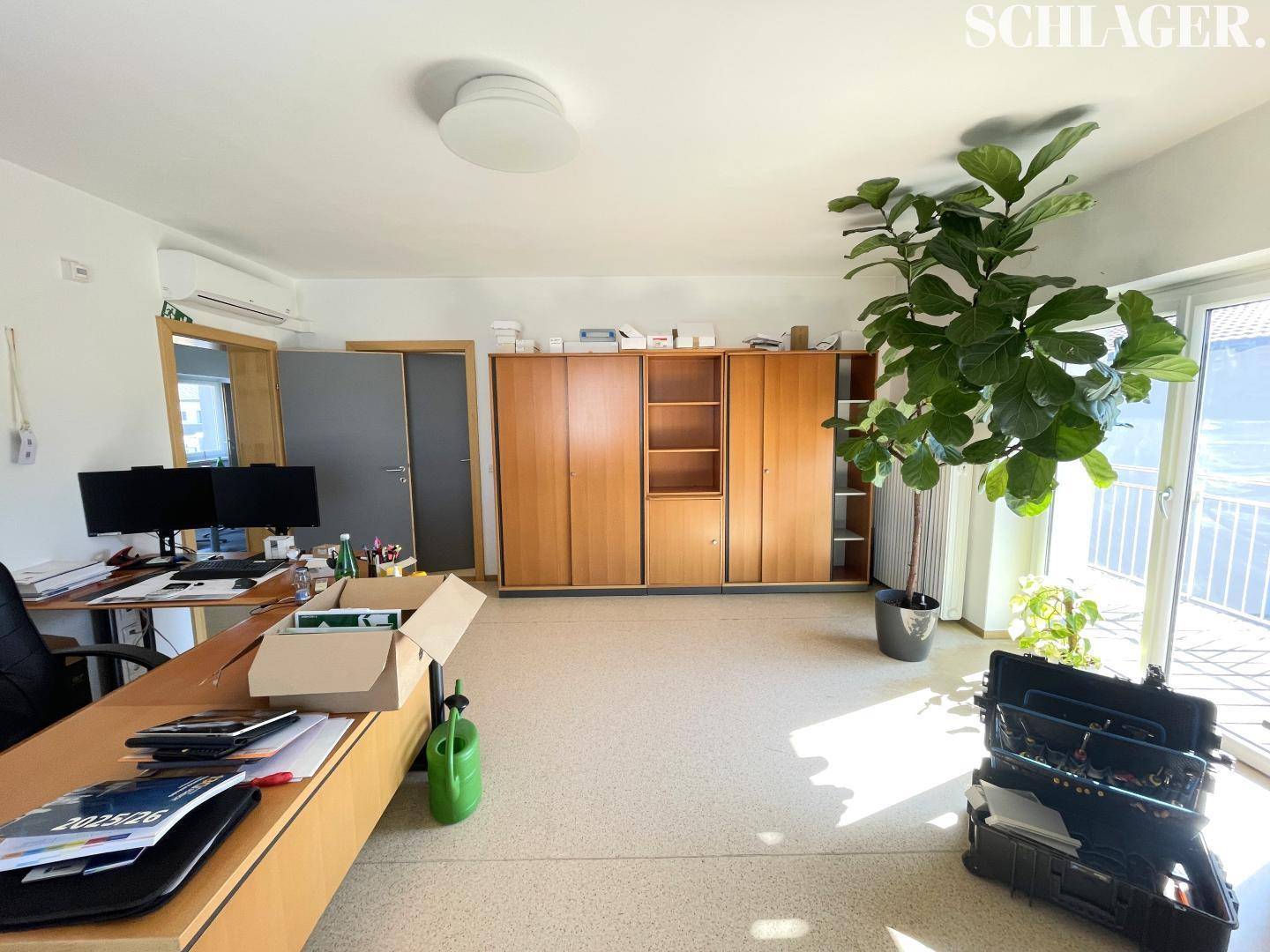 Geräumiges Büro mit Holzschränken, Schreibtisch und Zugang zu einem Balkon, hell durch große Fenster.