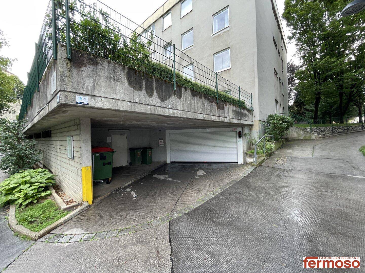 Zufahrt zur Tiefgarage mit einem Teil der Gebäudefassade und umgebenden Bäumen.