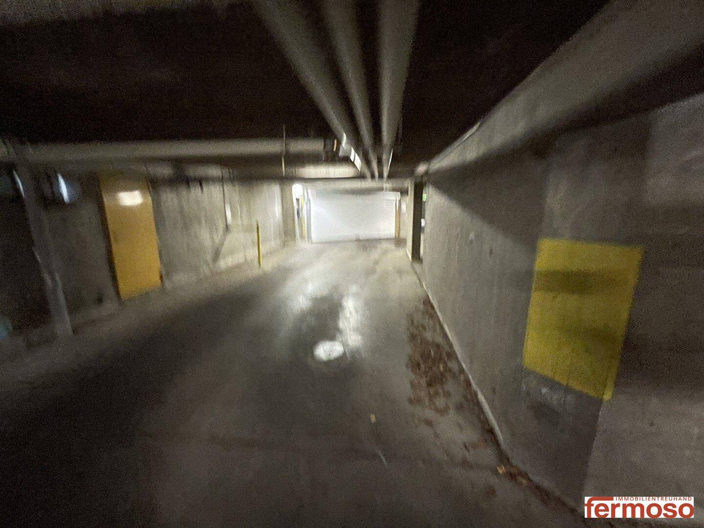 Langer Korridor in der Tiefgarage mit Betonwänden und sichtbaren Rohren an der Decke.
