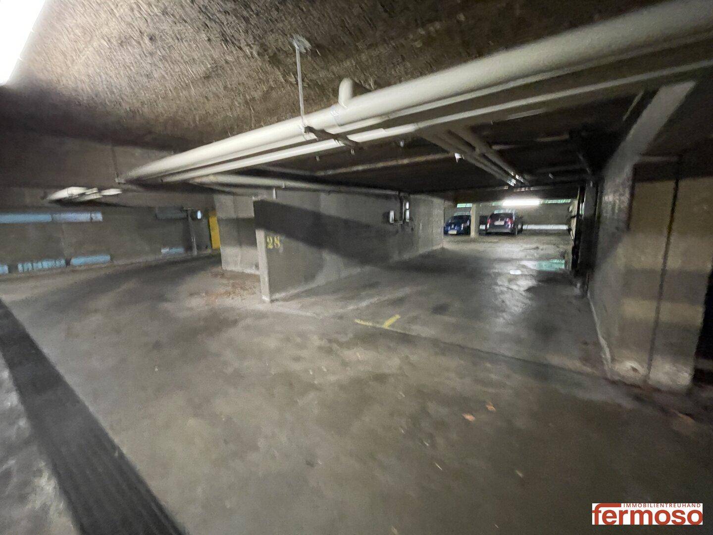 Weitläufige Tiefgarage mit vielen Stellplätzen und sichtbaren Versorgungsleitungen an der Decke.