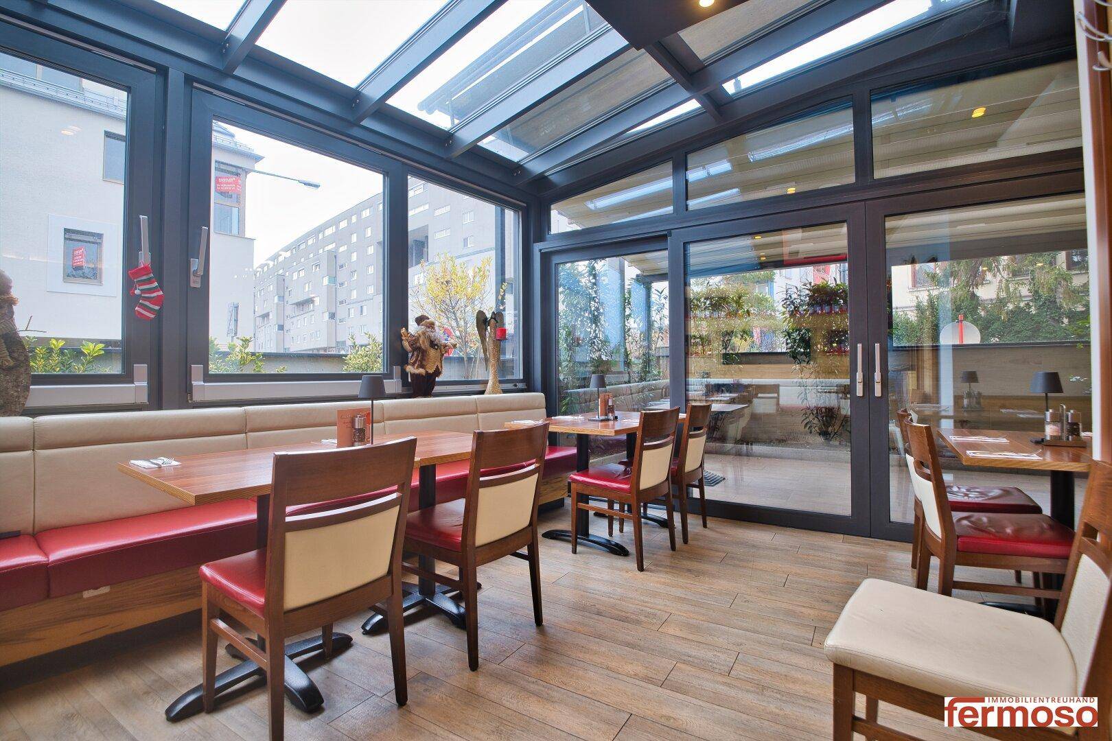 Heller Restaurantbereich in einem Wintergarten mit großen Glasfenstern und Holzböden.