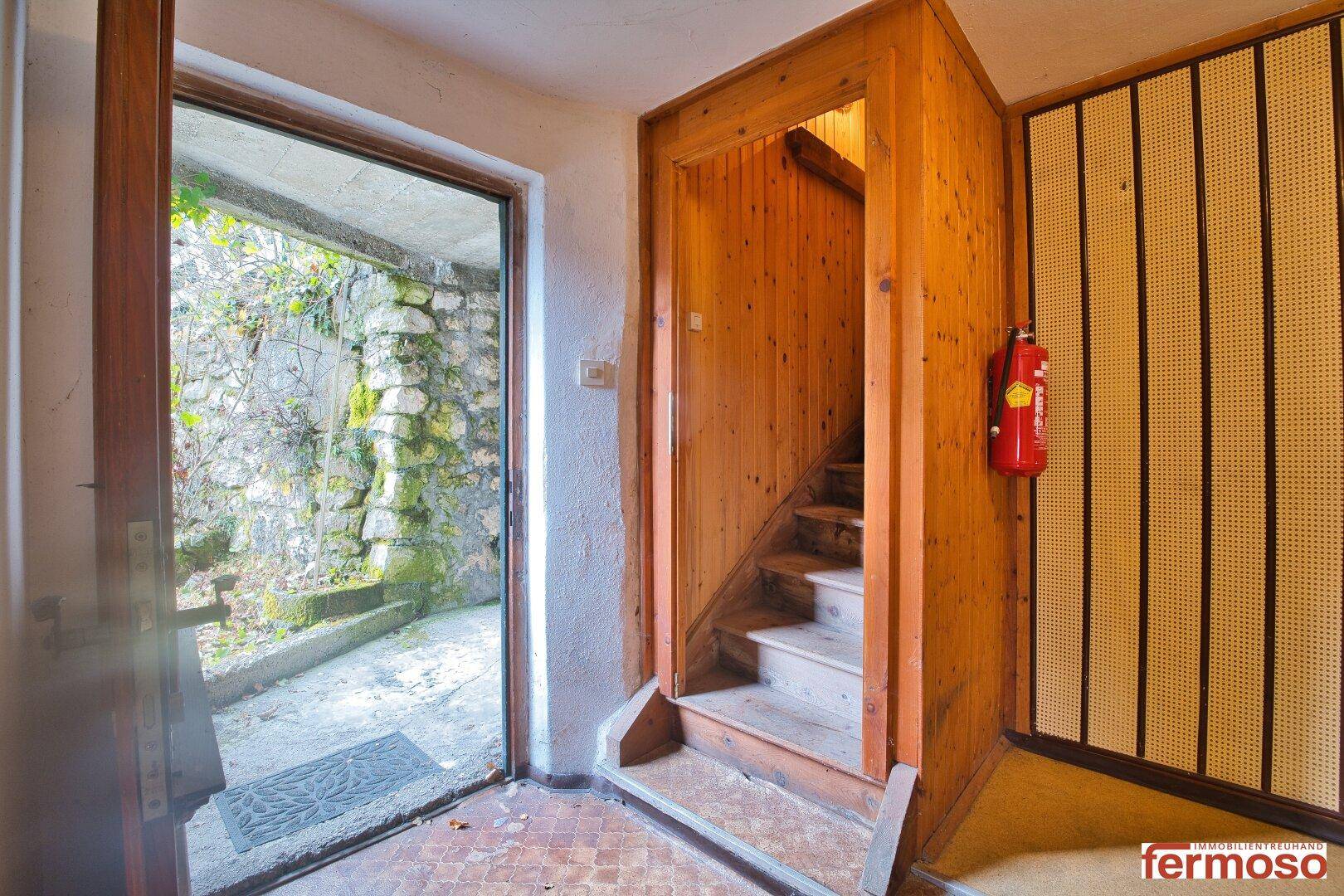 Eingangsbereich mit Holztreppe und Zugang zu einem begrünten Außenbereich mit Steinmauern.