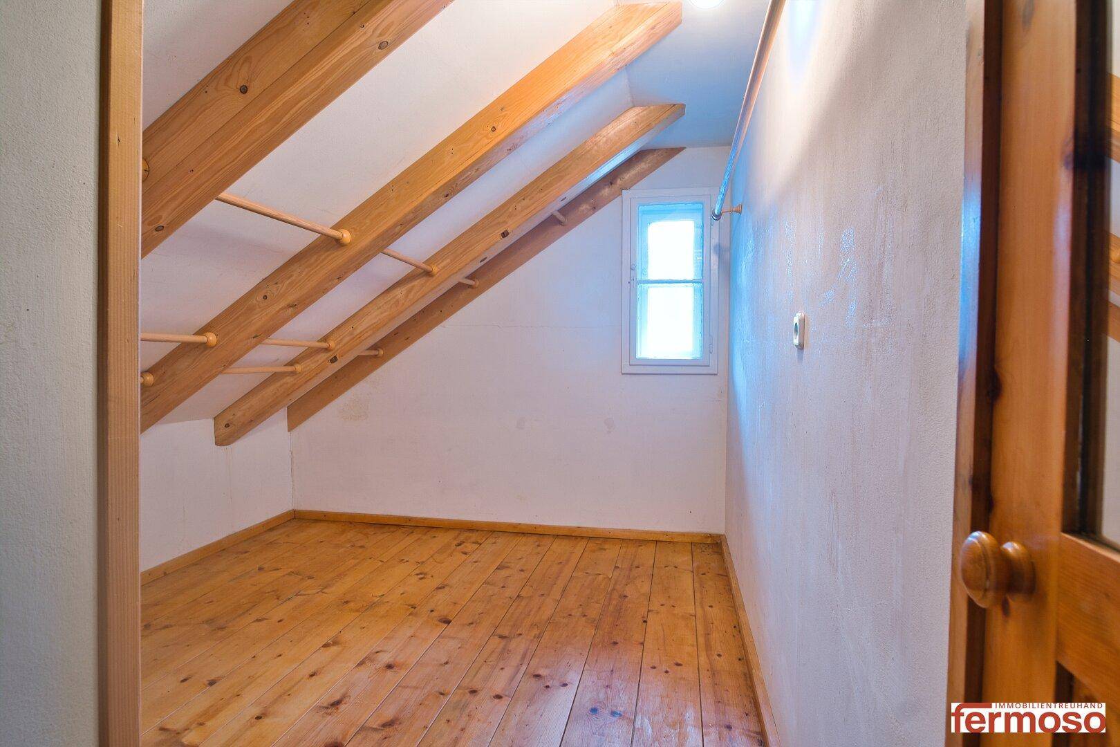 Leerer Dachbodenraum mit Holzboden, sichtbaren Balken und kleinem Fenster, hell und geräumig.