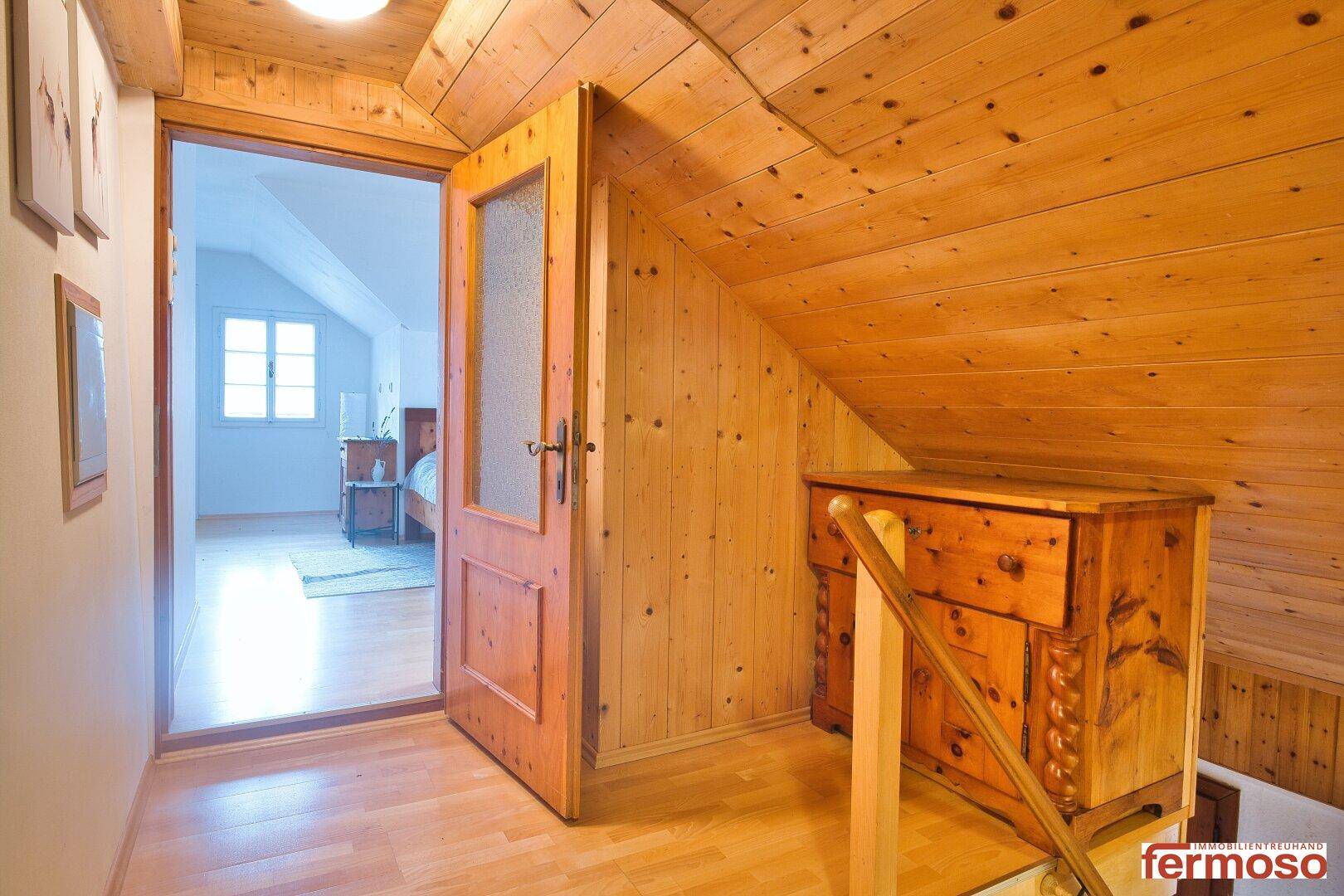 Dachgeschossflur mit Holzverkleidung, Holzboden und Zugang zu einem hellen Schlafzimmer.