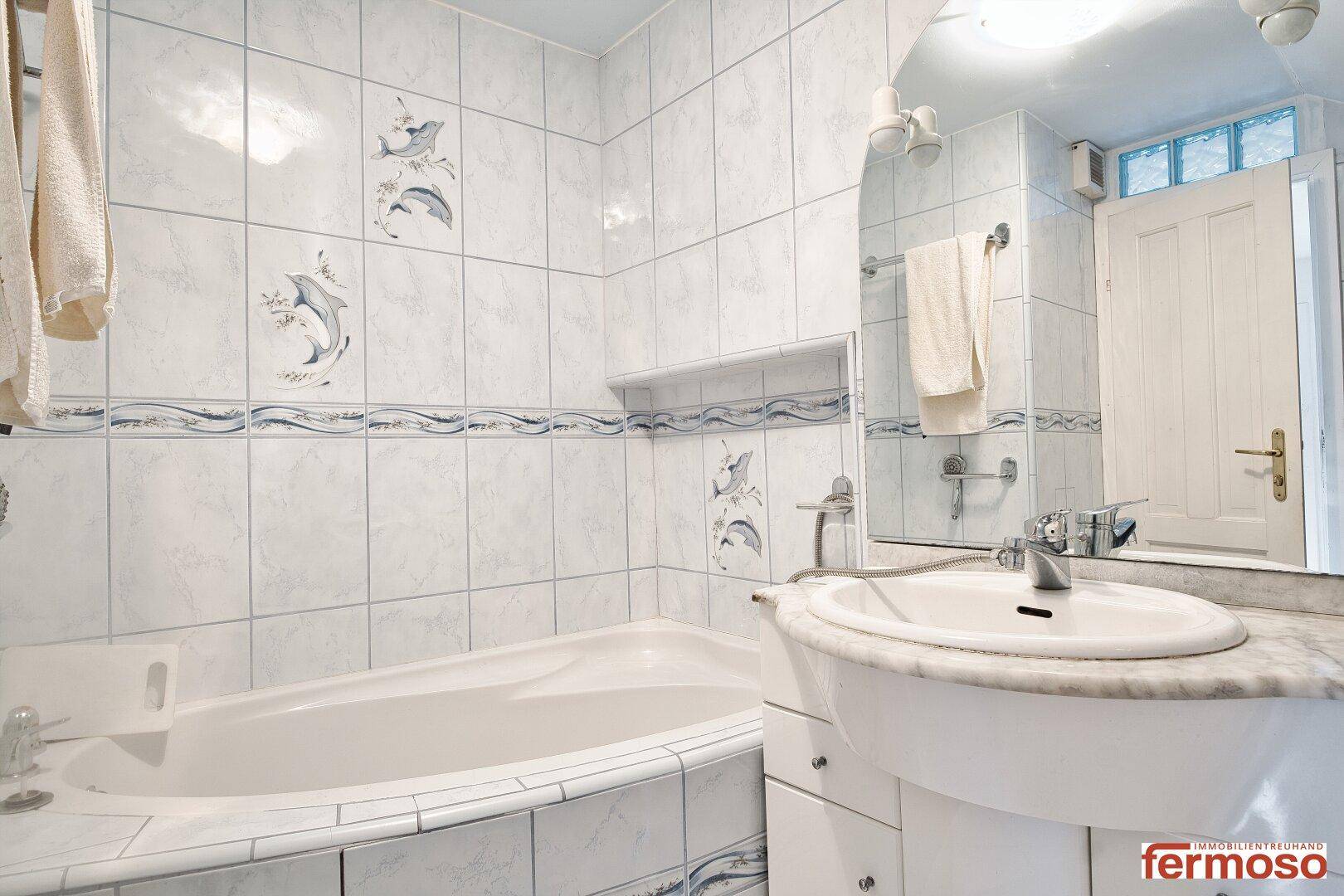 Helles Badezimmer mit Badewanne und Waschbecken, verziert mit blauen Fliesenbordüren und Delfinmotiven.