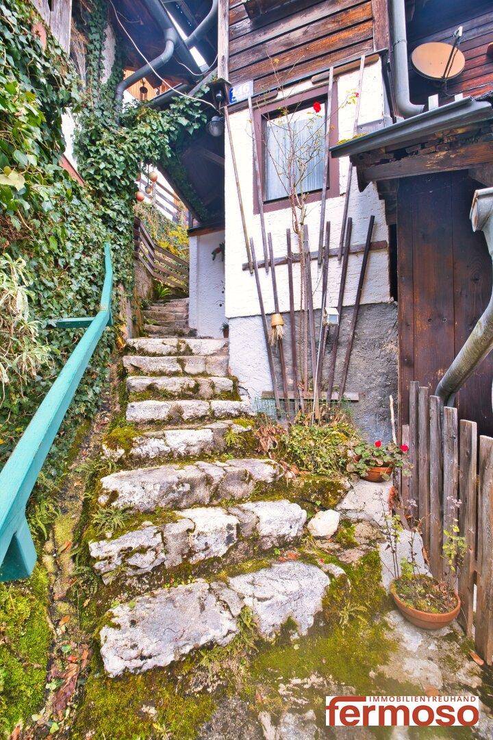 Moosbewachsene Steintreppe führt einen bewachsenen Hang hinauf, neben einem traditionellen Gebäude.