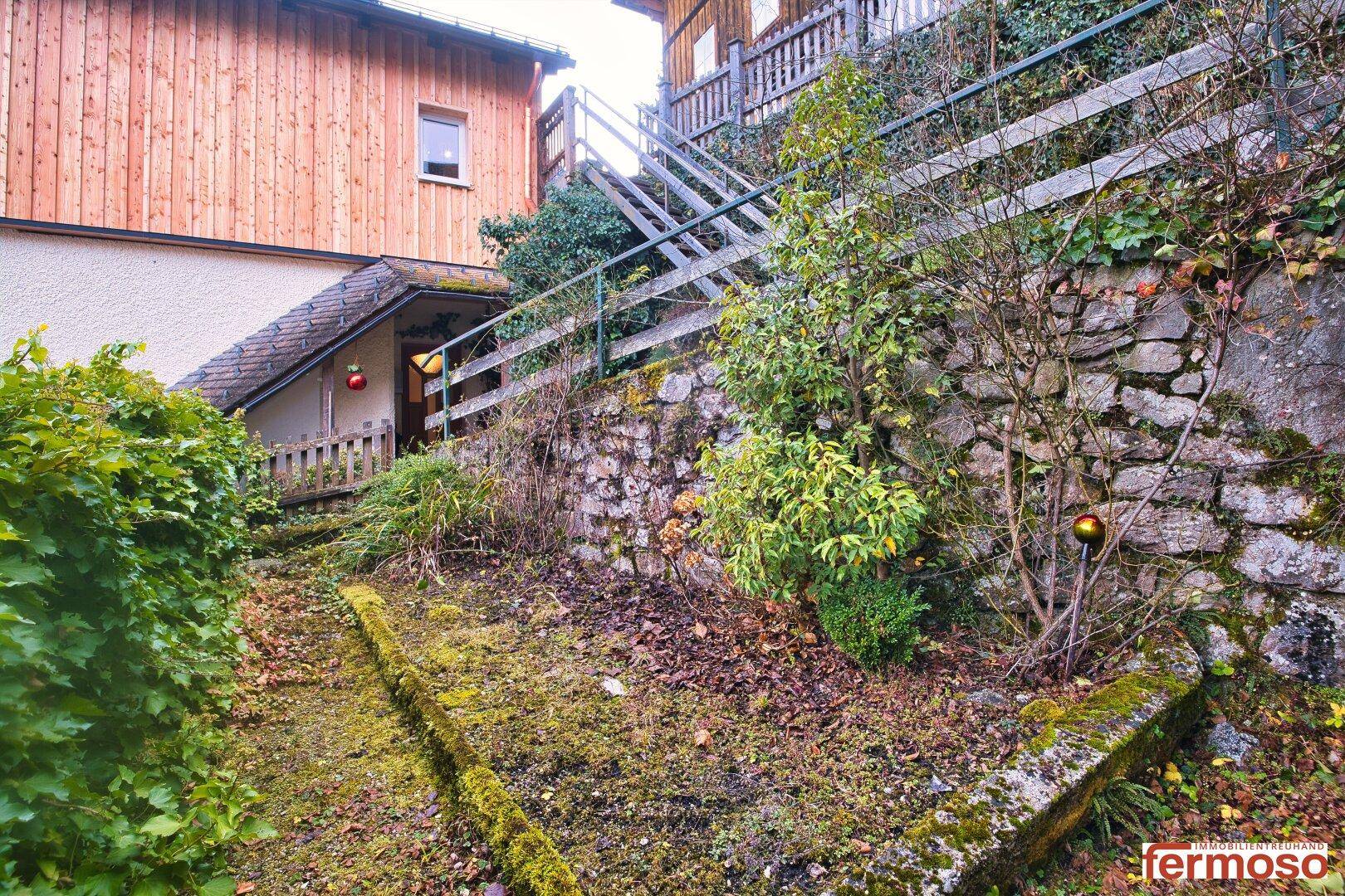 Bewachsener Hanggartenbereich mit Steinmauern und Stufen, angrenzend an ein traditionelles Haus.