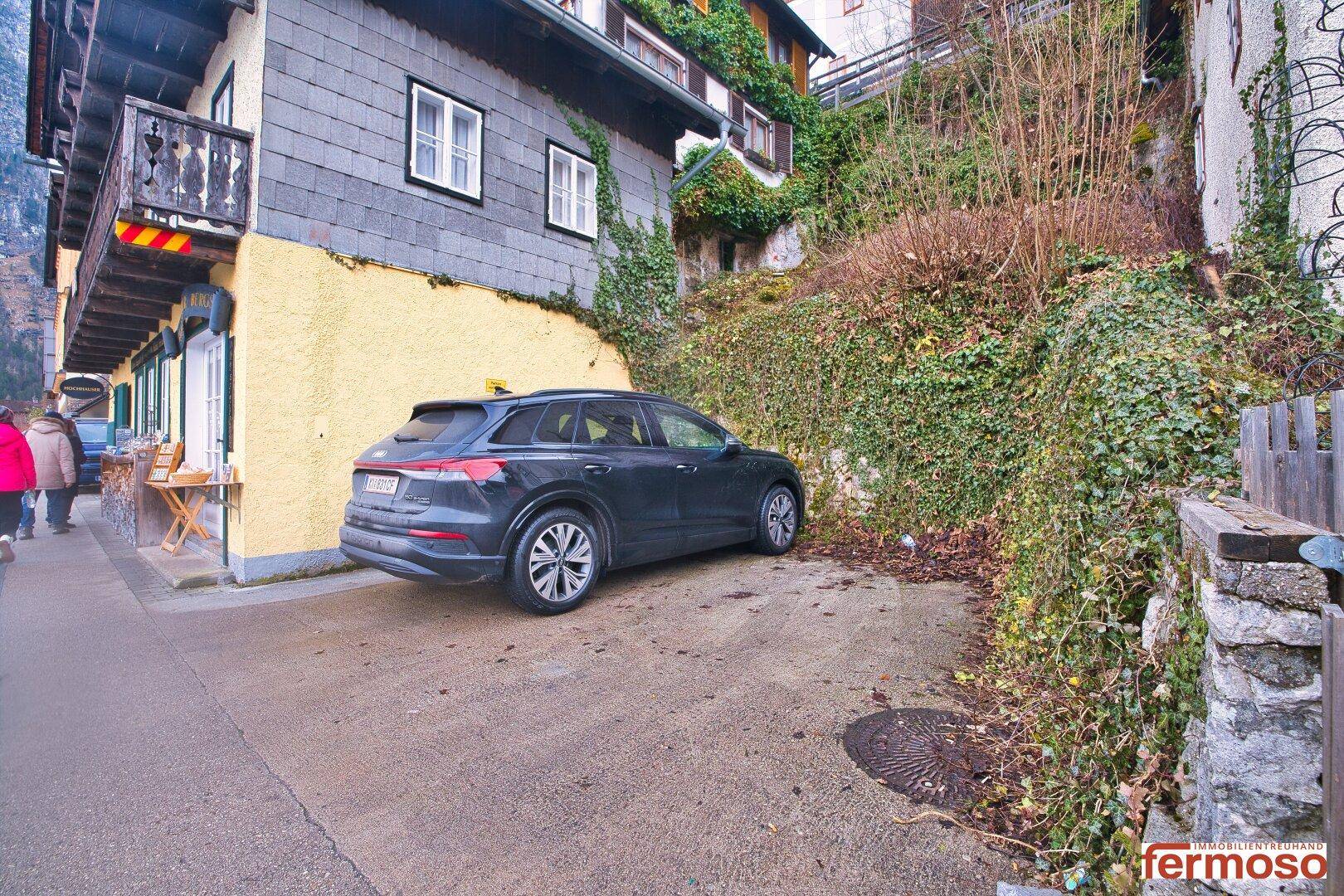 Parkplatz neben einem traditionellen Gebäude, umgeben von einem bewachsenen Hang.
