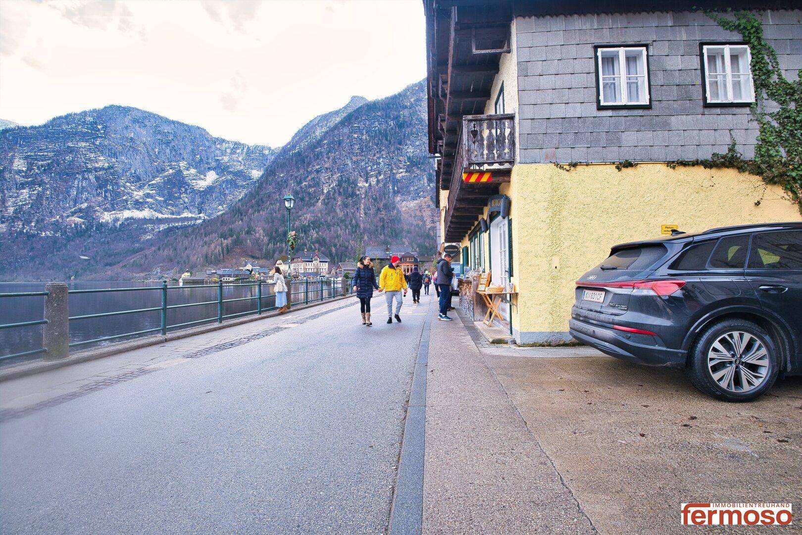 Straßenansicht entlang des Sees mit Spaziergängern, einem geparkten Auto und Bergen im Hintergrund.