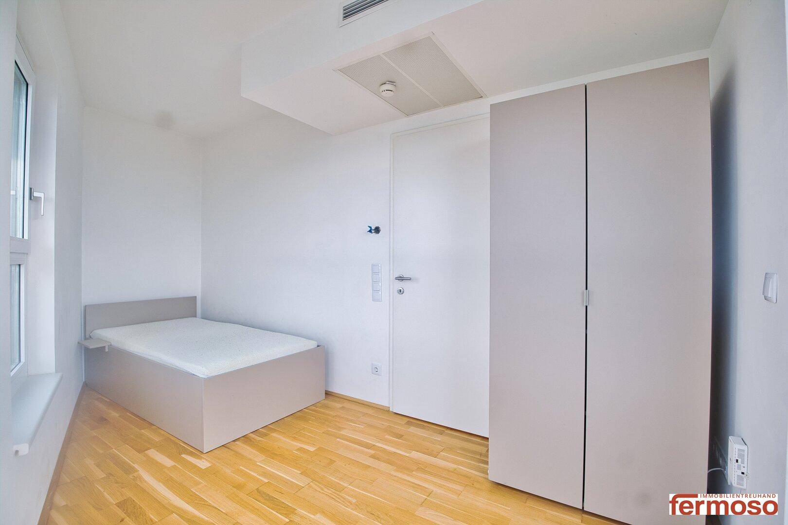 Helles Schlafzimmer mit einfachem Bettrahmen, modernem Kleiderschrank und Parkettboden.
