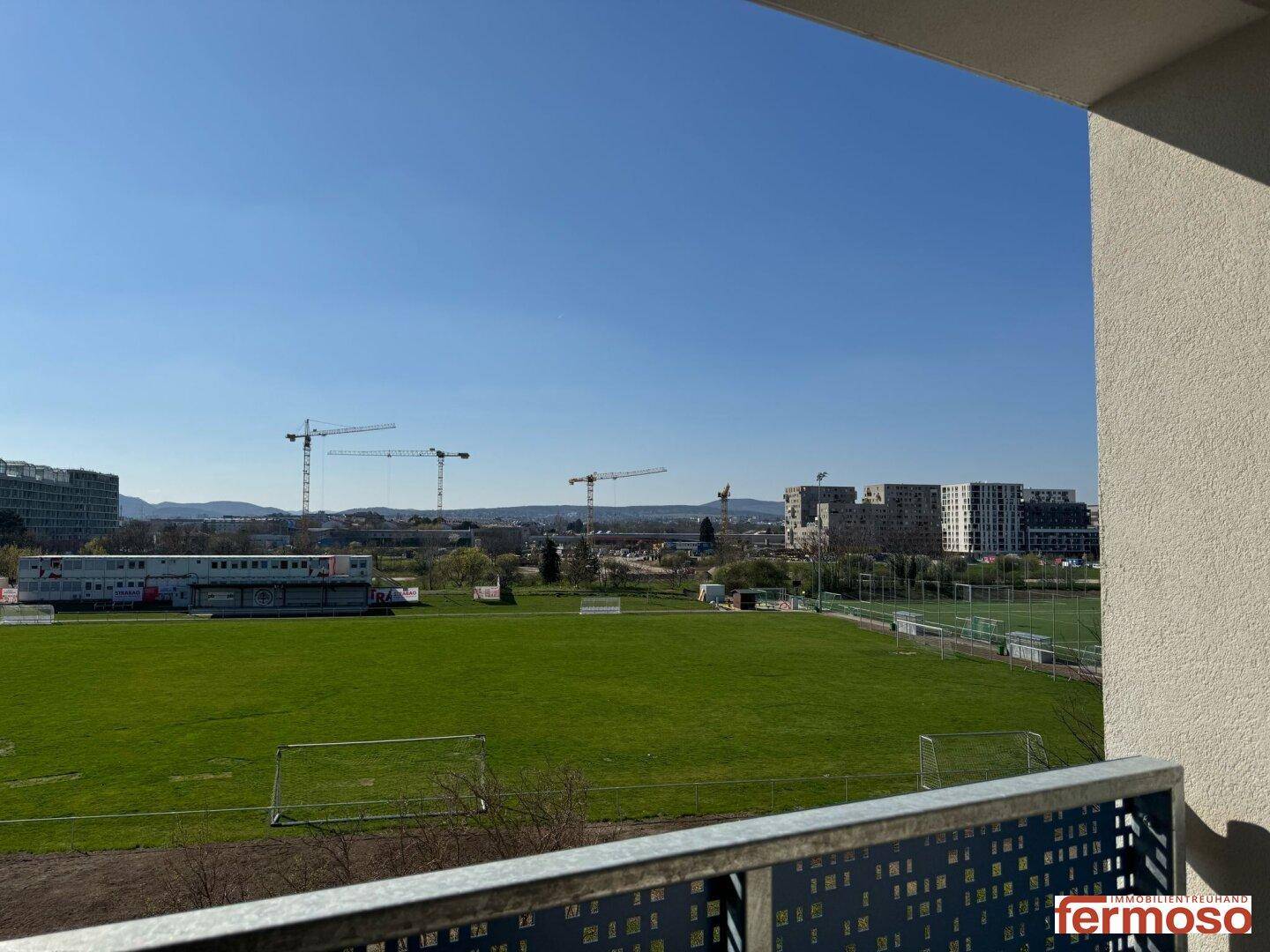Weitläufiger Blick von einem Balkon über ein grünes Feld und die städtische Skyline mit Baukränen.