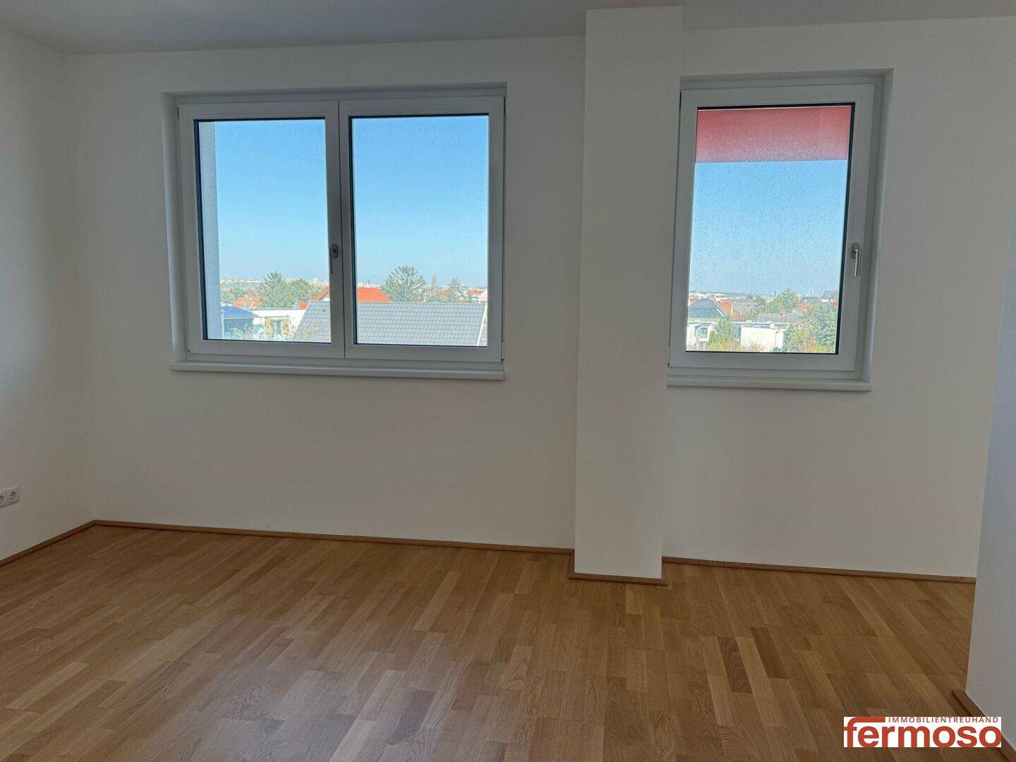 Leeres Zimmer mit hellem Parkettboden und zwei Fenstern, die einen weiten Ausblick bieten.